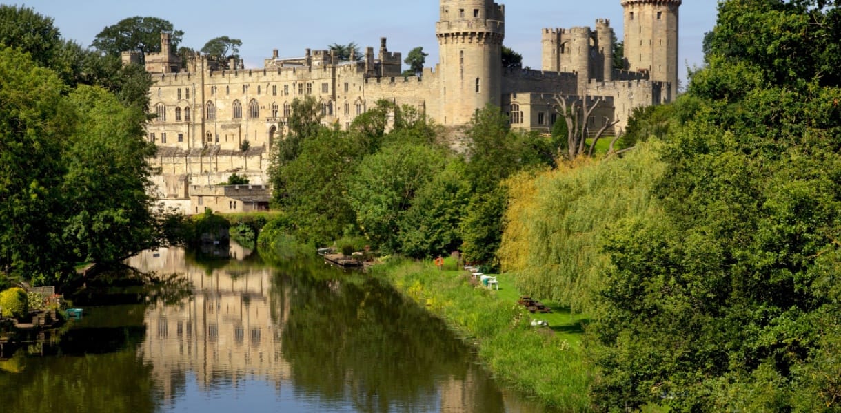 View of Warwick Castle, in Warwickshire, rising above the River Avon, surrounded by lush green trees and reflected in the calm water below, with clear blue skies in the background