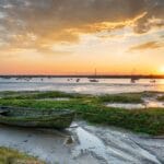 Sunset over the salt marshes at West Mersea in Essex, with a weathered wooden boat in the foreground, calm water dotted with moored sailing boats, and a dramatic sky reflecting golden hues across the estuary.