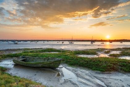 Sunset over the salt marshes at West Mersea in Essex, with a weathered wooden boat in the foreground, calm water dotted with moored sailing boats, and a dramatic sky reflecting golden hues across the estuary.