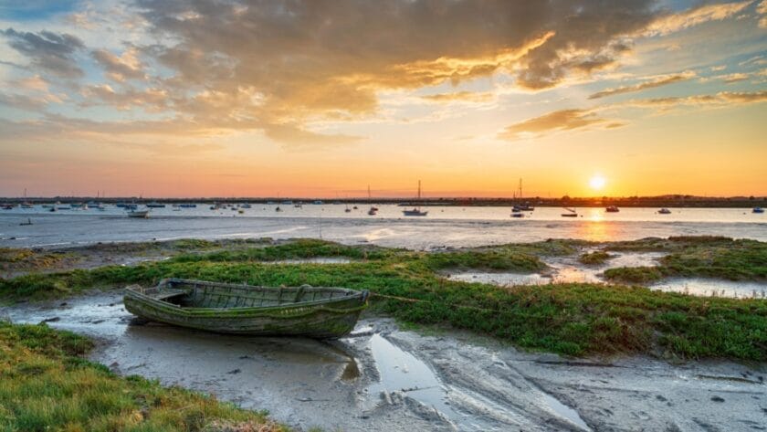 Sunset over the salt marshes at West Mersea in Essex, with a weathered wooden boat in the foreground, calm water dotted with moored sailing boats, and a dramatic sky reflecting golden hues across the estuary.