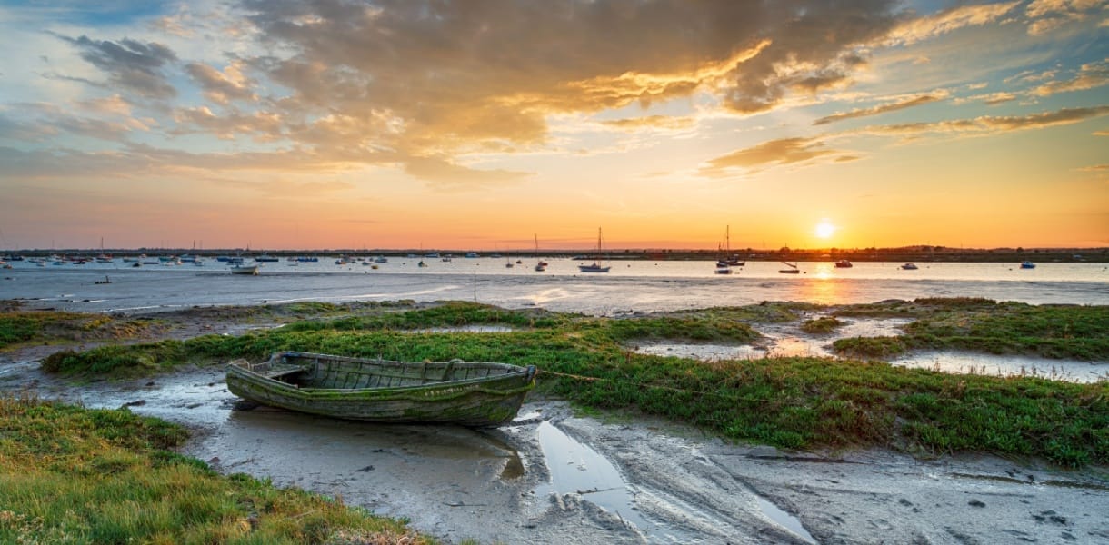 Sunset over the salt marshes at West Mersea in Essex, with a weathered wooden boat in the foreground, calm water dotted with moored sailing boats, and a dramatic sky reflecting golden hues across the estuary.