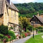 Charming honey-coloured stone cottages with flower-filled gardens line a quiet street in Broadway, a picturesque village in the Cotswolds area of Worcestershire, England, with lush green hills in the background.