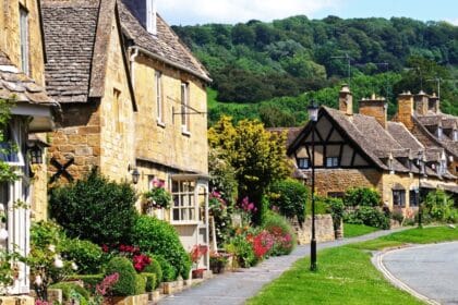 Charming honey-coloured stone cottages with flower-filled gardens line a quiet street in Broadway, a picturesque village in the Cotswolds area of Worcestershire, England, with lush green hills in the background.