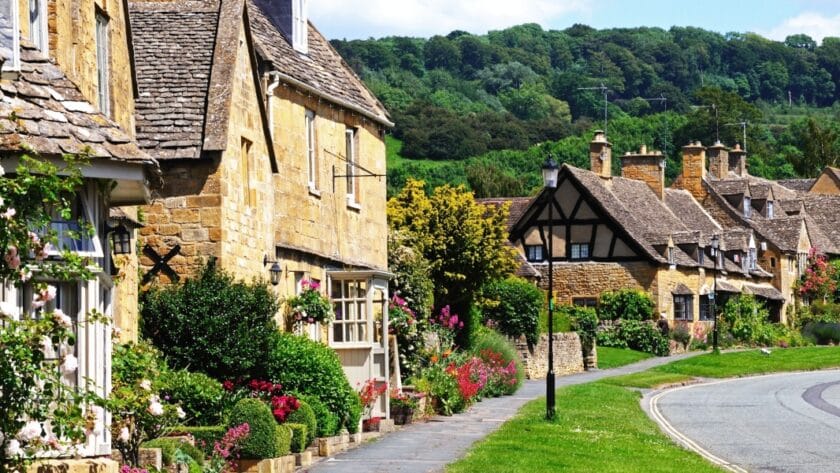 Charming honey-coloured stone cottages with flower-filled gardens line a quiet street in Broadway, a picturesque village in the Cotswolds area of Worcestershire, England, with lush green hills in the background.