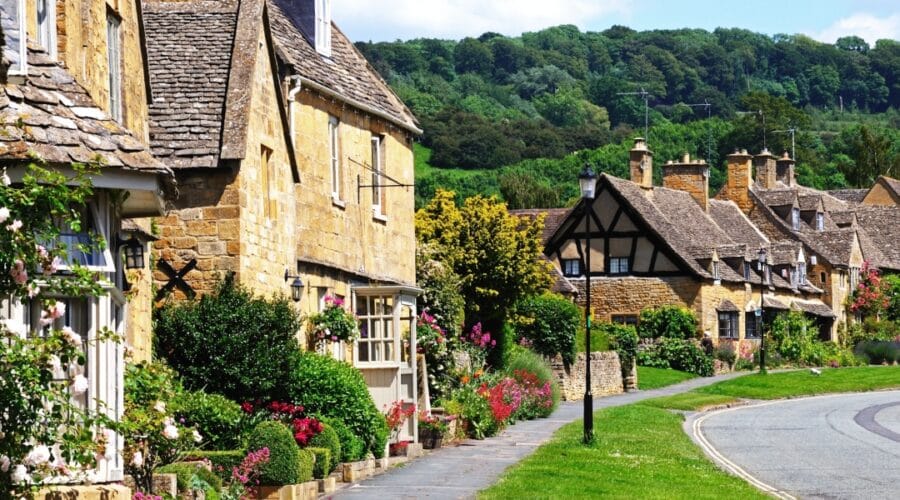 Charming honey-coloured stone cottages with flower-filled gardens line a quiet street in Broadway, a picturesque village in the Cotswolds area of Worcestershire, England, with lush green hills in the background.