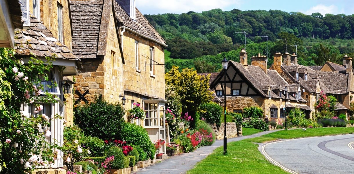 Charming honey-coloured stone cottages with flower-filled gardens line a quiet street in Broadway, a picturesque village in the Cotswolds area of Worcestershire, England, with lush green hills in the background.
