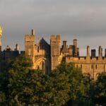 Arundel Castle in West Sussex, its stone towers and battlements glowing in warm evening light, rising above a line of green trees under a moody sky