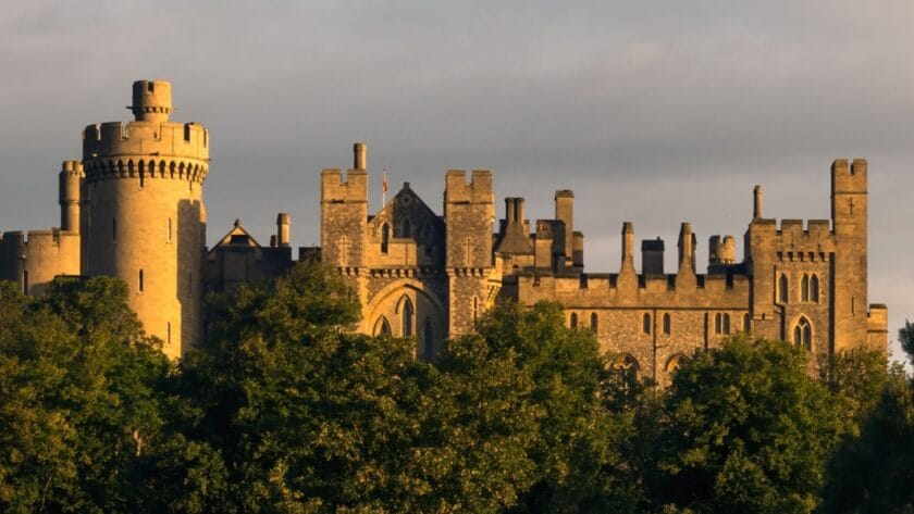 Arundel Castle in West Sussex, its stone towers and battlements glowing in warm evening light, rising above a line of green trees under a moody sky