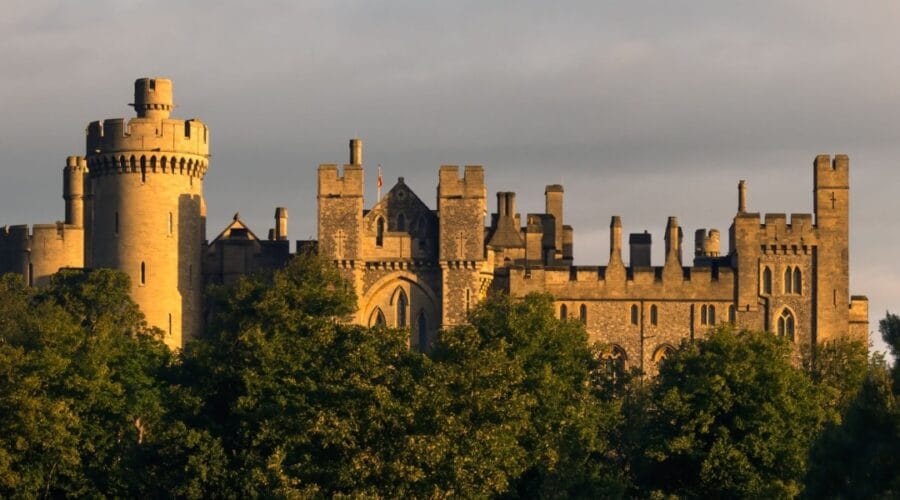 Arundel Castle in West Sussex, its stone towers and battlements glowing in warm evening light, rising above a line of green trees under a moody sky