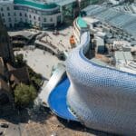 Aerial view of central Birmingham showing the modern Selfridges building with its curved silver façade covered in circular discs beside the historic St Martin’s Church, surrounded by shoppers and city streets