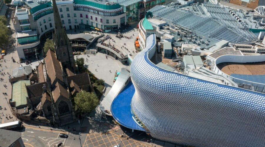 Aerial view of central Birmingham showing the modern Selfridges building with its curved silver façade covered in circular discs beside the historic St Martin’s Church, surrounded by shoppers and city streets