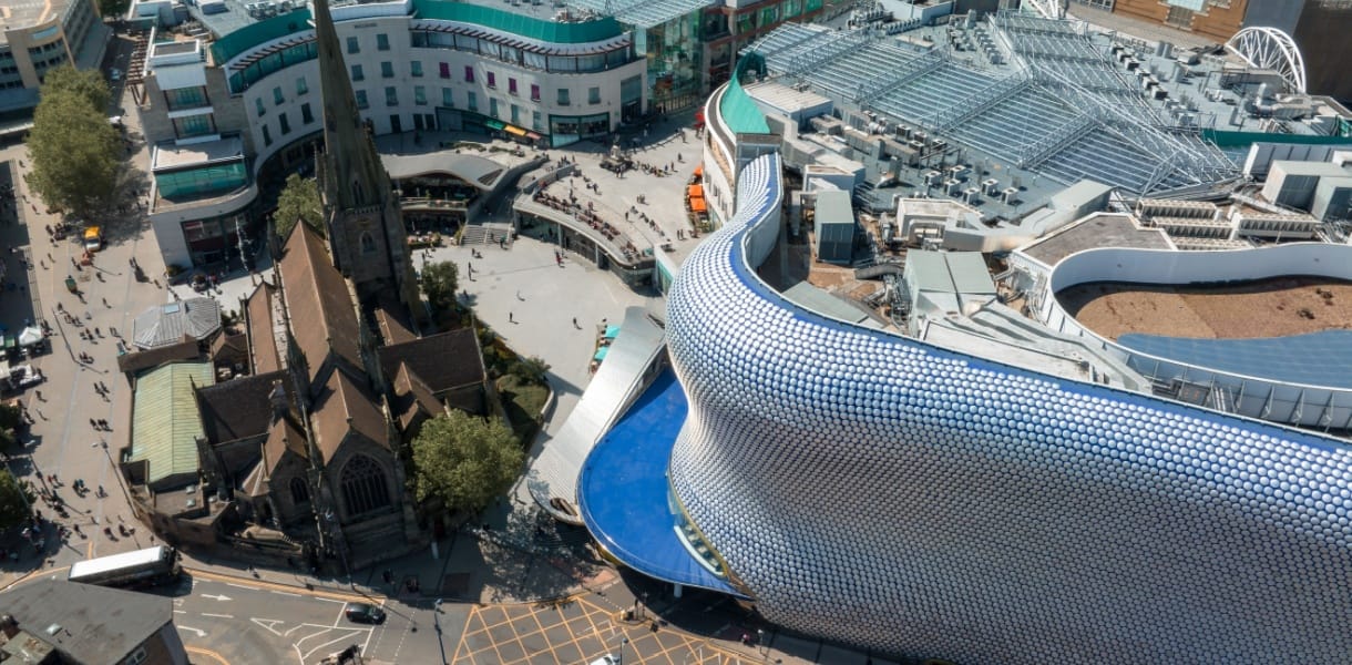 Aerial view of central Birmingham showing the modern Selfridges building with its curved silver façade covered in circular discs beside the historic St Martin’s Church, surrounded by shoppers and city streets