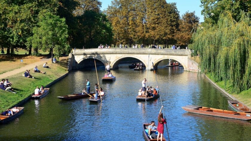 Visitors punting on the River Cam in Cambridge, gliding past the historic Clare Bridge with students and tourists relaxing on the riverbanks, a classic Cambridge weekend experience.