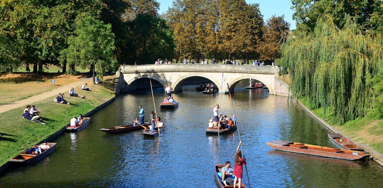Visitors punting on the River Cam in Cambridge, gliding past the historic Clare Bridge with students and tourists relaxing on the riverbanks, a classic Cambridge weekend experience.