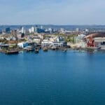 A panoramic view of Cardiff Bay with a sailboat in the foreground, the waterfront lined with modern buildings, the red-brick Pierhead Building, the Wales Millennium Centre, and a large Ferris wheel, with the city skyline and stadium visible in the background under a clear blue sky