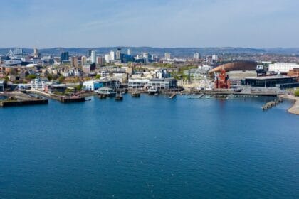 A panoramic view of Cardiff Bay with a sailboat in the foreground, the waterfront lined with modern buildings, the red-brick Pierhead Building, the Wales Millennium Centre, and a large Ferris wheel, with the city skyline and stadium visible in the background under a clear blue sky