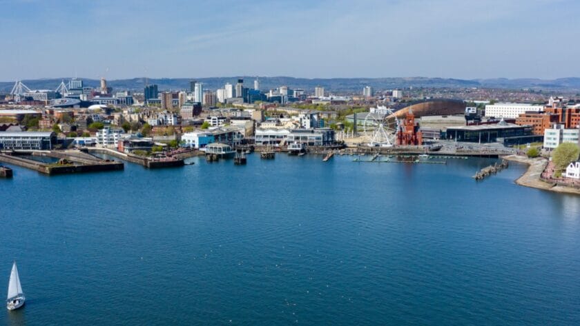 A panoramic view of Cardiff Bay with a sailboat in the foreground, the waterfront lined with modern buildings, the red-brick Pierhead Building, the Wales Millennium Centre, and a large Ferris wheel, with the city skyline and stadium visible in the background under a clear blue sky