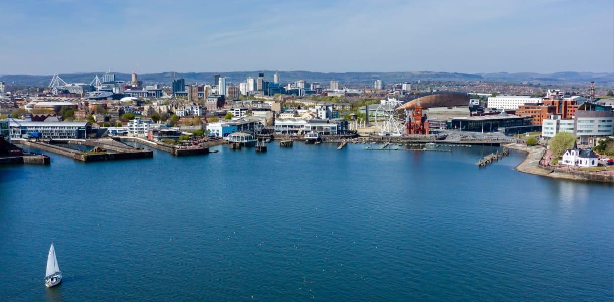 A panoramic view of Cardiff Bay with a sailboat in the foreground, the waterfront lined with modern buildings, the red-brick Pierhead Building, the Wales Millennium Centre, and a large Ferris wheel, with the city skyline and stadium visible in the background under a clear blue sky