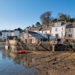 Waterside cottages in Fowey, Cornwall, with stone steps leading down to the riverbank, a small red boat floating on calm water, and wooded hills rising in the background under a clear blue sky