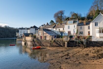 Waterside cottages in Fowey, Cornwall, with stone steps leading down to the riverbank, a small red boat floating on calm water, and wooded hills rising in the background under a clear blue sky