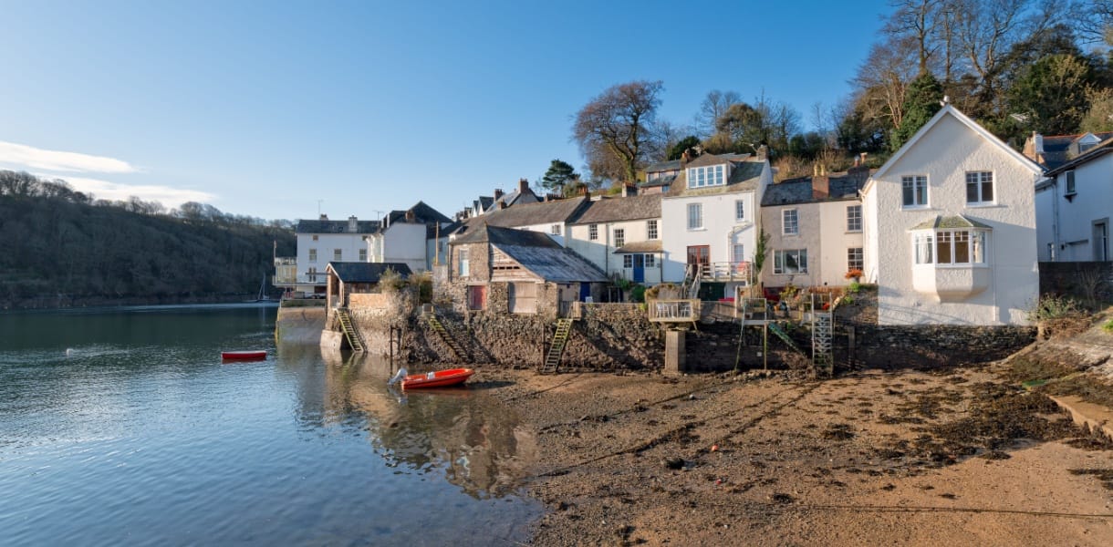 Waterside cottages in Fowey, Cornwall, with stone steps leading down to the riverbank, a small red boat floating on calm water, and wooded hills rising in the background under a clear blue sky