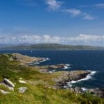 Rocky coastline of the Isle of Bute with green hillsides in the foreground, waves breaking along the shore, and a sailboat gliding across the deep blue waters of the Firth of Clyde under a bright sky with scattered clouds.