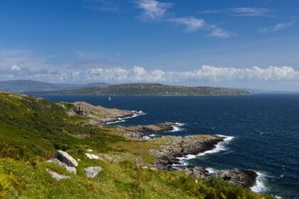 Rocky coastline of the Isle of Bute with green hillsides in the foreground, waves breaking along the shore, and a sailboat gliding across the deep blue waters of the Firth of Clyde under a bright sky with scattered clouds.