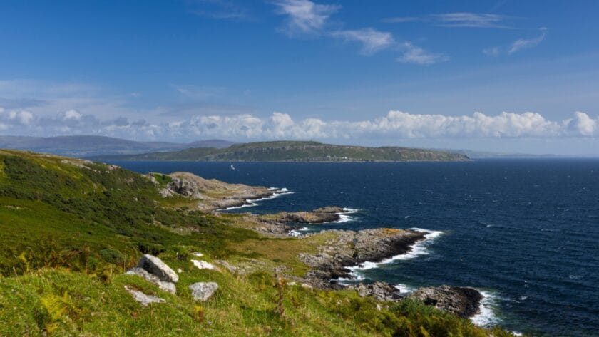 Rocky coastline of the Isle of Bute with green hillsides in the foreground, waves breaking along the shore, and a sailboat gliding across the deep blue waters of the Firth of Clyde under a bright sky with scattered clouds.