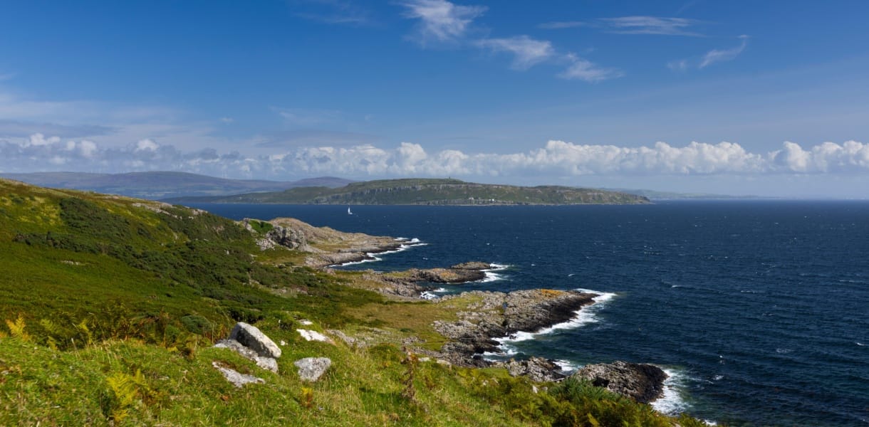 Rocky coastline of the Isle of Bute with green hillsides in the foreground, waves breaking along the shore, and a sailboat gliding across the deep blue waters of the Firth of Clyde under a bright sky with scattered clouds.
