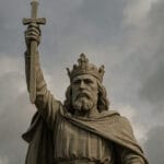 A stone statue of King Alfred the Great stands against a cloudy sky, depicting the Anglo-Saxon king wearing a crown and cloak. His right arm is raised high, holding a cross-topped sword, with a stormy sky in the background