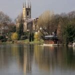 View of Kingston upon Thames from the riverside, with the River Thames in the foreground reflecting trees and boats moored along the bank. The historic All Saints Church with its tall clock tower rises above the town, framed by leafy greenery and riverside buildings under a soft, cloudy sky