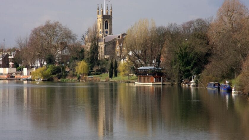 View of Kingston upon Thames from the riverside, with the River Thames in the foreground reflecting trees and boats moored along the bank. The historic All Saints Church with its tall clock tower rises above the town, framed by leafy greenery and riverside buildings under a soft, cloudy sky