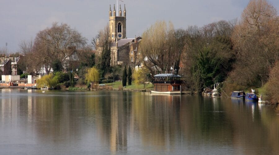 View of Kingston upon Thames from the riverside, with the River Thames in the foreground reflecting trees and boats moored along the bank. The historic All Saints Church with its tall clock tower rises above the town, framed by leafy greenery and riverside buildings under a soft, cloudy sky