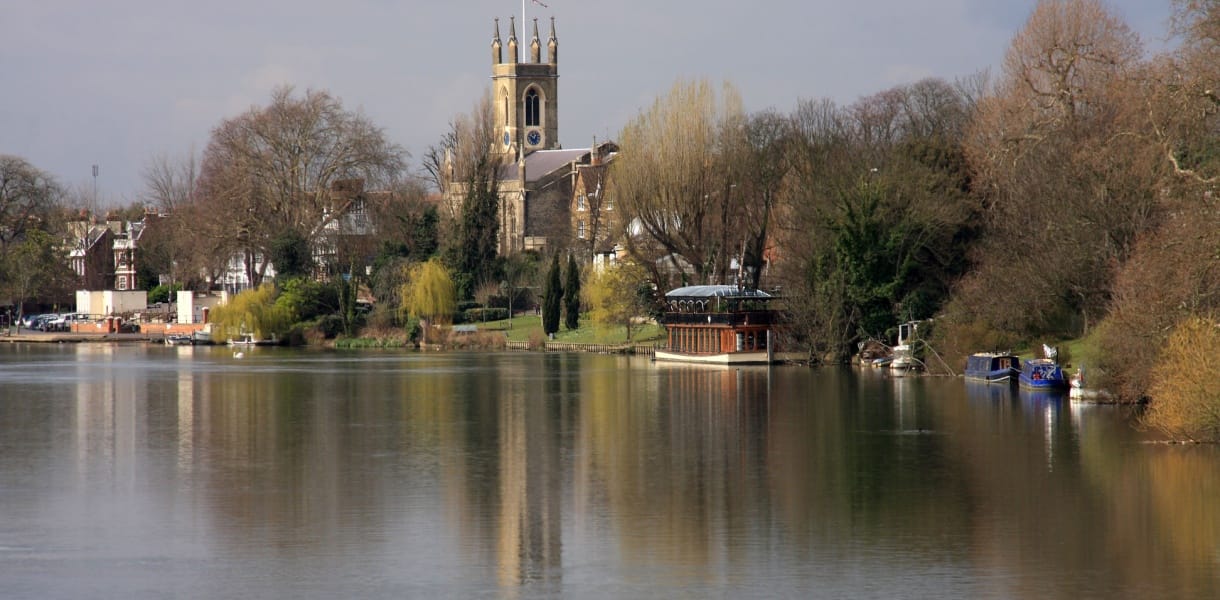 View of Kingston upon Thames from the riverside, with the River Thames in the foreground reflecting trees and boats moored along the bank. The historic All Saints Church with its tall clock tower rises above the town, framed by leafy greenery and riverside buildings under a soft, cloudy sky