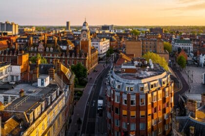 Aerial view of Leicester city centre at sunset, showing historic red-brick buildings, the clock tower, and modern blocks in the distance, with golden light illuminating the rooftops and streets