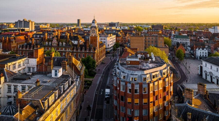Aerial view of Leicester city centre at sunset, showing historic red-brick buildings, the clock tower, and modern blocks in the distance, with golden light illuminating the rooftops and streets