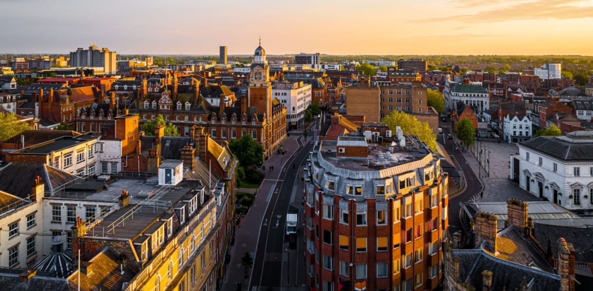 Aerial view of Leicester city centre at sunset, showing historic red-brick buildings, the clock tower, and modern blocks in the distance, with golden light illuminating the rooftops and streets