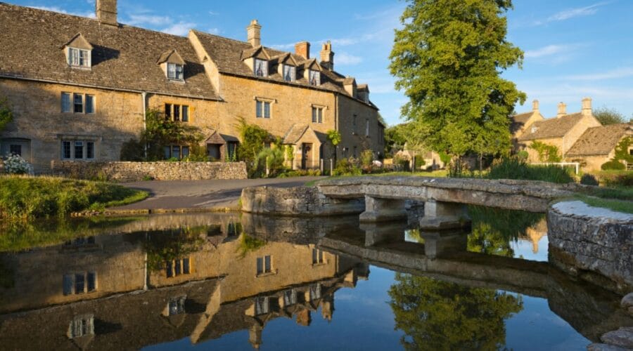 Scenic view of Lower Slaughter in the Cotswolds, featuring honey-coloured stone cottages, a tree-lined stream, and a small stone footbridge reflected in the water