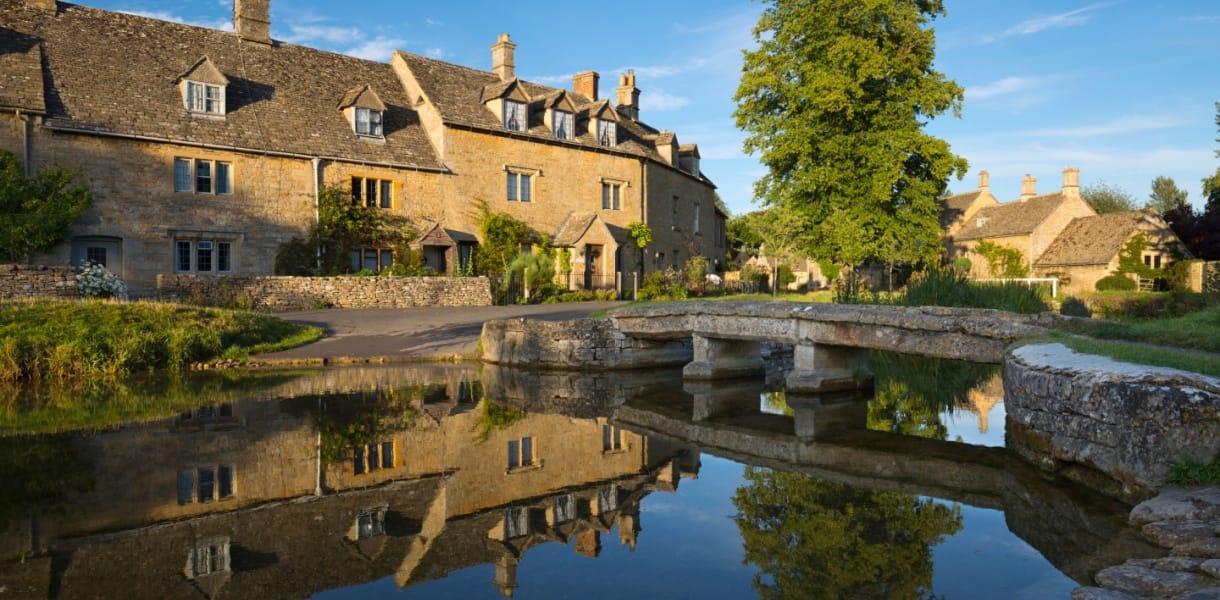 Scenic view of Lower Slaughter in the Cotswolds, featuring honey-coloured stone cottages, a tree-lined stream, and a small stone footbridge reflected in the water
