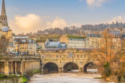 View of Pulteney Bridge in Bath, England, spanning the River Avon with its elegant Georgian architecture. The bridge is lined with historic stone buildings, and the surrounding cityscape features honey-coloured terraces, a church spire, and rolling hills in the background under a soft, partly cloudy sky