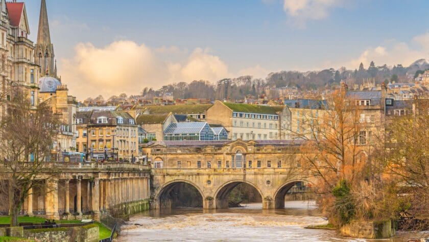 View of Pulteney Bridge in Bath, England, spanning the River Avon with its elegant Georgian architecture. The bridge is lined with historic stone buildings, and the surrounding cityscape features honey-coloured terraces, a church spire, and rolling hills in the background under a soft, partly cloudy sky