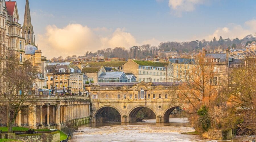 View of Pulteney Bridge in Bath, England, spanning the River Avon with its elegant Georgian architecture. The bridge is lined with historic stone buildings, and the surrounding cityscape features honey-coloured terraces, a church spire, and rolling hills in the background under a soft, partly cloudy sky