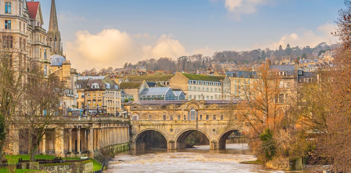 View of Pulteney Bridge in Bath, England, spanning the River Avon with its elegant Georgian architecture. The bridge is lined with historic stone buildings, and the surrounding cityscape features honey-coloured terraces, a church spire, and rolling hills in the background under a soft, partly cloudy sky