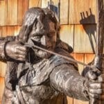 Close-up of a bronze statue of Robin Hood drawing his bow, with a quiver of arrows on his back, set against a backdrop of wooden shingles