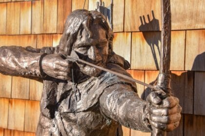 Close-up of a bronze statue of Robin Hood drawing his bow, with a quiver of arrows on his back, set against a backdrop of wooden shingles