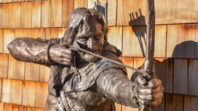 Close-up of a bronze statue of Robin Hood drawing his bow, with a quiver of arrows on his back, set against a backdrop of wooden shingles