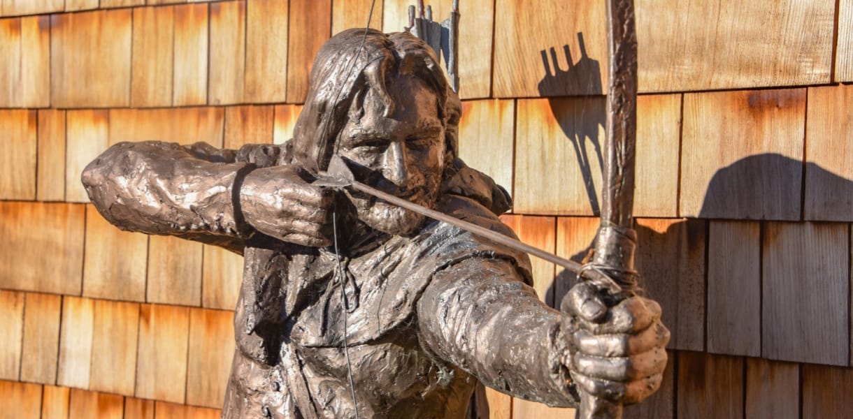 Close-up of a bronze statue of Robin Hood drawing his bow, with a quiver of arrows on his back, set against a backdrop of wooden shingles