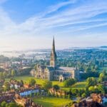 Aerial view of Salisbury Cathedral in Wiltshire, with its soaring spire rising above surrounding trees, historic red-roofed houses, and misty countryside under a bright blue sky