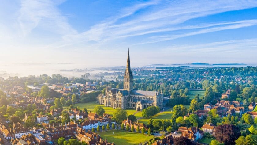 Aerial view of Salisbury Cathedral in Wiltshire, with its soaring spire rising above surrounding trees, historic red-roofed houses, and misty countryside under a bright blue sky
