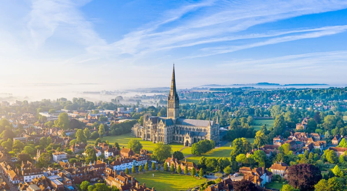 Aerial view of Salisbury Cathedral in Wiltshire, with its soaring spire rising above surrounding trees, historic red-roofed houses, and misty countryside under a bright blue sky
